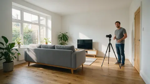 A bright living room with a grey sofa and plants, a man with a camera on a tripod at the side
