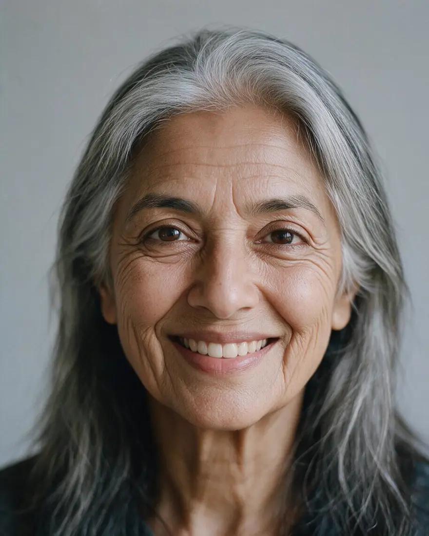 About 70: Age this woman to about 70 years old: visible wrinkles, mostly grey hair in the same long style. Keep exactly the same warm medium-brown skin tone as the photo — do not lighten the skin. Keep her identity, face shape, eye shape, smile, expression, clothing, the background and lighting the same.