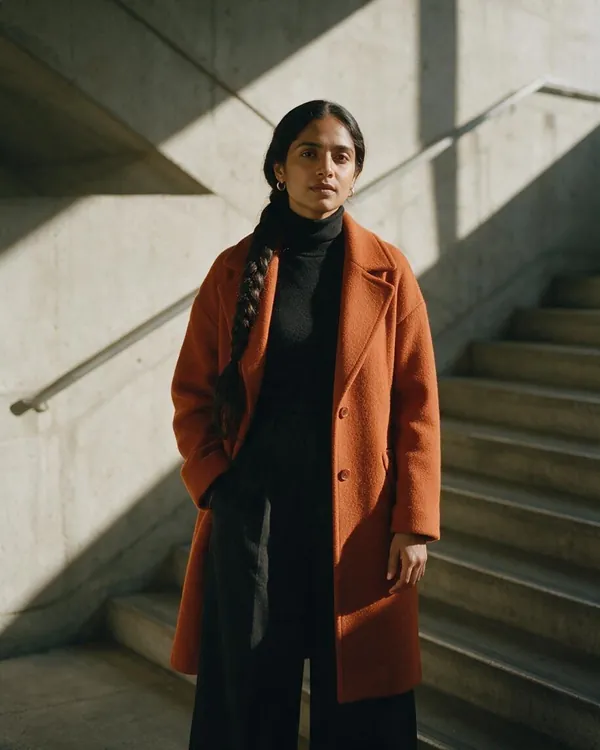 AI-generated editorial photo of a woman with a long dark braid in a rust-orange wool coat and black turtleneck standing in a sunlit concrete stairwell