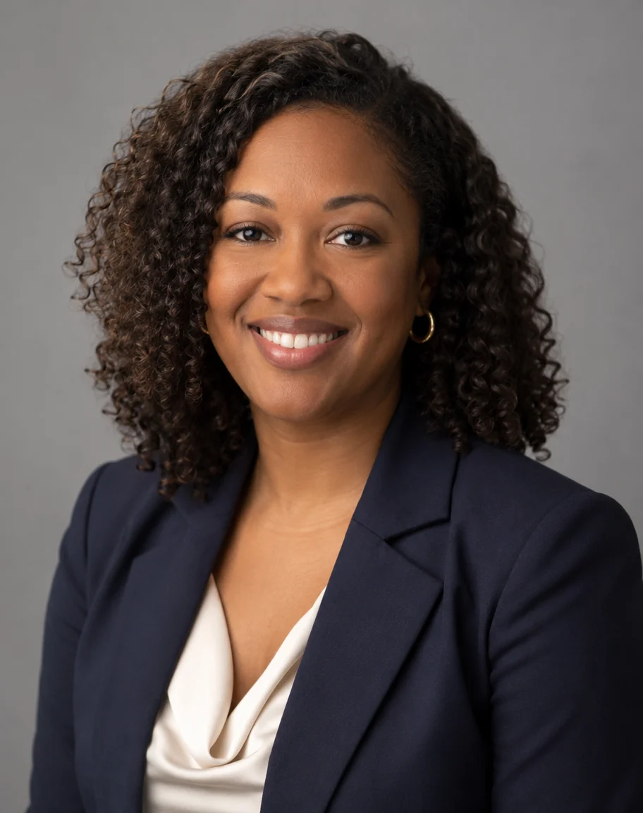 AI-generated studio headshot of a smiling woman in her late 30s with curly shoulder-length hair, a navy blazer and a cream blouse on a grey background