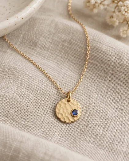 Hammered yellow gold disc pendant with a small bezel-set blue sapphire on a fine chain, laid on beige linen