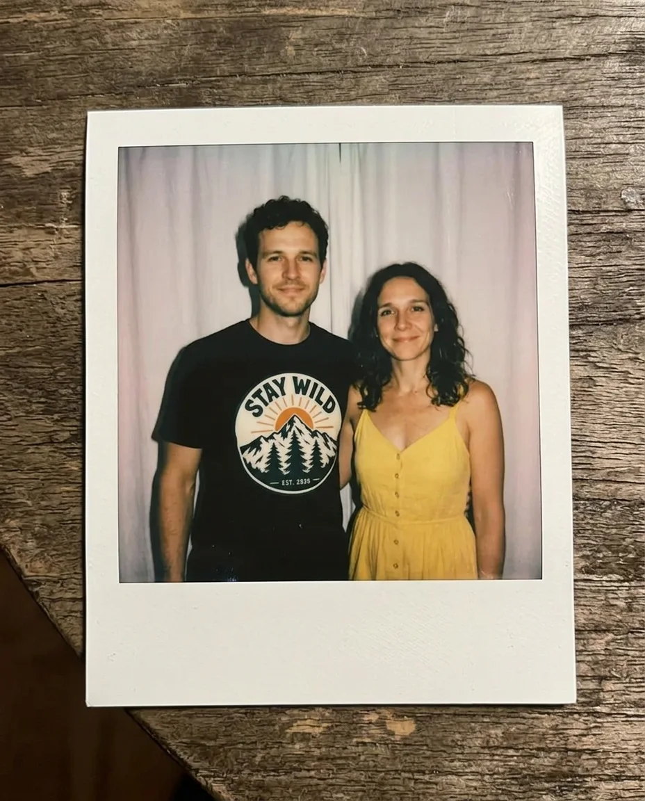 A Polaroid print on a wooden table showing a curly-haired man in a STAY WILD t-shirt and a woman in a yellow sundress standing in front of a white curtain