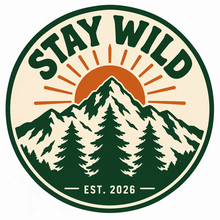 A circular STAY WILD t-shirt badge with a mountain, three pine trees and a rising sun in green, orange and cream