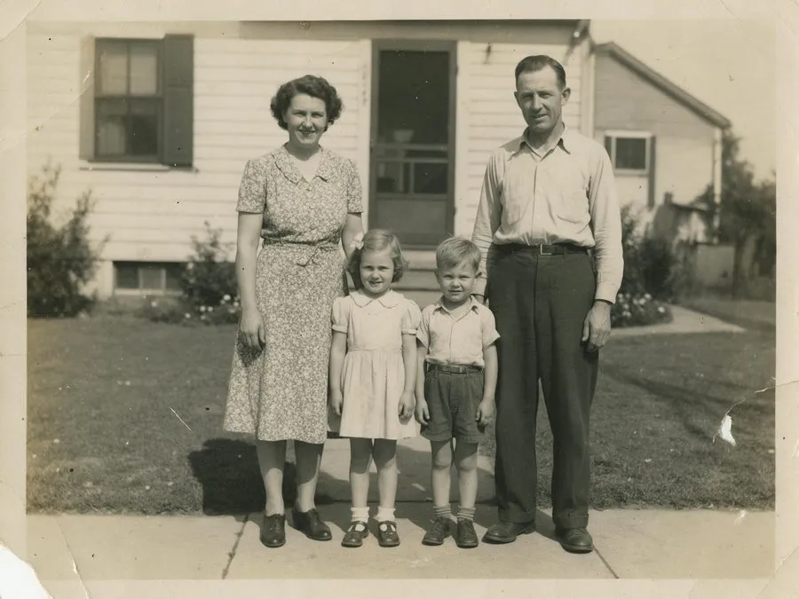 Faded 1950s black-and-white family photo in front of a house, with scratches and a torn corner — before